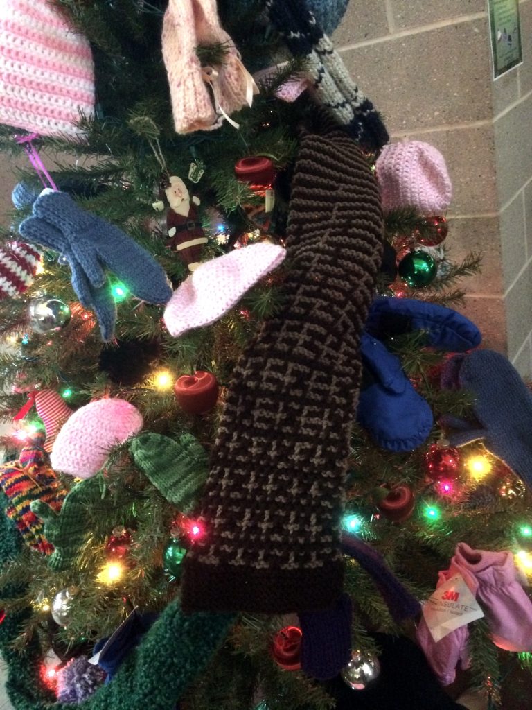 A picture of a Christmas Tree decorated with mittens, gloves, hats and scarves as well as lights and ornaments